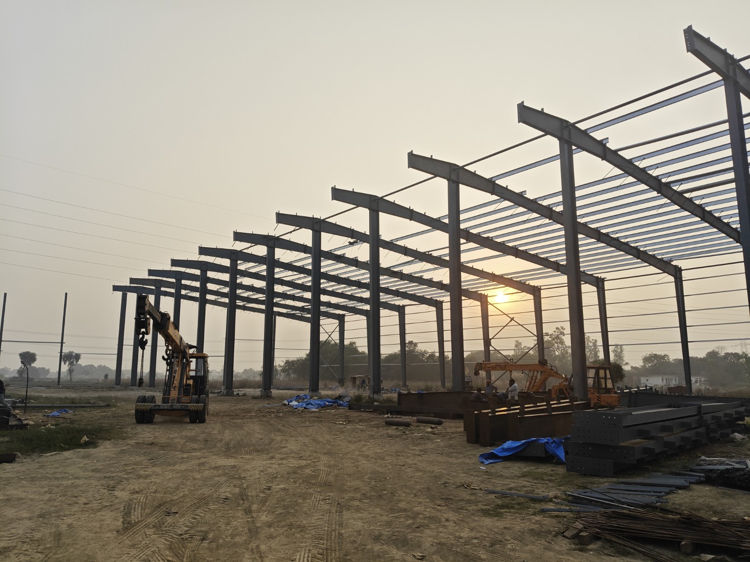 Steel columns, rafters and roof purlins during erection, with site lifting equipment at sunset.